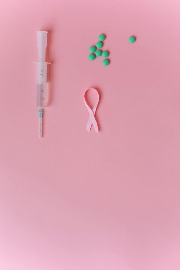 Flat lay of a syringe, pink ribbon, and green pills on a soft pink background symbolizing breast cancer awareness.