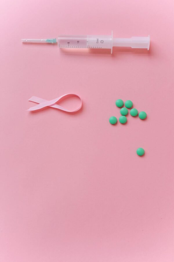 Pink ribbon, syringe, and green pills on pink background symbolizing breast cancer awareness and health care.