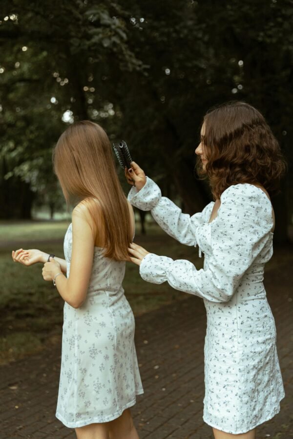 Two women in floral dresses combing hair in a serene park, emphasizing friendship and care.