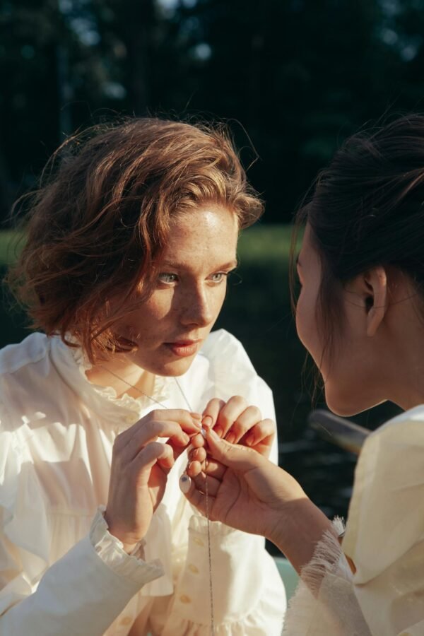 Two women sit at a riverside, gently examining a delicate necklace in the sunlight.